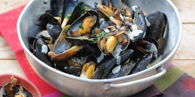 Moules marinières aux amandes dans une cocotte en métal Moules cuisinées dans une cocotte en métal, ouvertes et garnies d'amandes effilées, d'oignons et d'herbes, sur une table en bois.